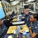 A group of U.S. Navy personnel in digital camouflage (NWU Type I) uniforms are gathered in what appears to be a ship's mess hall or wardroom. A senior officer is engaged in conversation with enlisted sailors seated at tables with food trays. The setting includes nautical charts, plaques on the walls, and an American flag in the background.