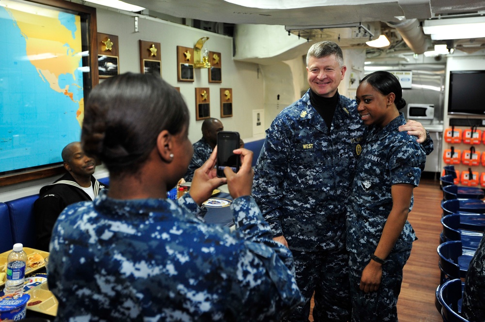 A group of U.S. Navy personnel are gathered in what appears to be a shipboard common area or mess deck. A sailor is photographing two colleagues — one identified by name tape as 'WEST' — who are posing together for a photo. The setting includes naval awards plaques, a world map, and standard shipboard furnishings.