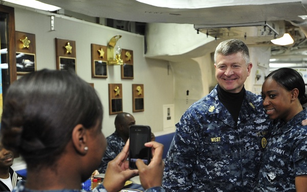 A group of U.S. Navy personnel are gathered in what appears to be a shipboard common area or mess deck. A sailor is photographing two colleagues — one identified by name tape as 'WEST' — who are posing together for a photo. The setting includes naval awards plaques, a world map, and standard shipboard furnishings.