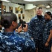 A group of U.S. Navy personnel are gathered in what appears to be a shipboard common area or mess deck. A sailor is photographing two colleagues — one identified by name tape as 'WEST' — who are posing together for a photo. The setting includes naval awards plaques, a world map, and standard shipboard furnishings.