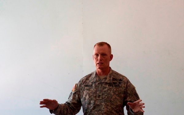 A U.S. Army soldier, identified by name tape as 'ARNOLD,' is addressing a group of seated military personnel in what appears to be a briefing or training session. He is standing at the front of a room with a plain white wall behind him, gesturing with both hands as he speaks. The audience, visible from behind, appears to include soldiers in camouflage uniforms.
