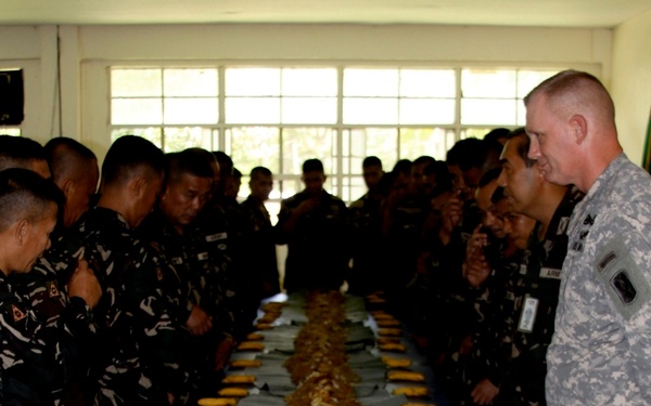 A large group of military personnel in two different uniforms — Philippine Armed Forces camouflage and US Army ACU (Army Combat Uniform) — are gathered around a long table covered with a blue tablecloth. The table features a traditional Filipino 'boodle fight' feast, with food served on banana leaves running the length of the table, accompanied by bananas placed at intervals. The group appears to be in a pre-meal prayer or moment of reflection before eating.