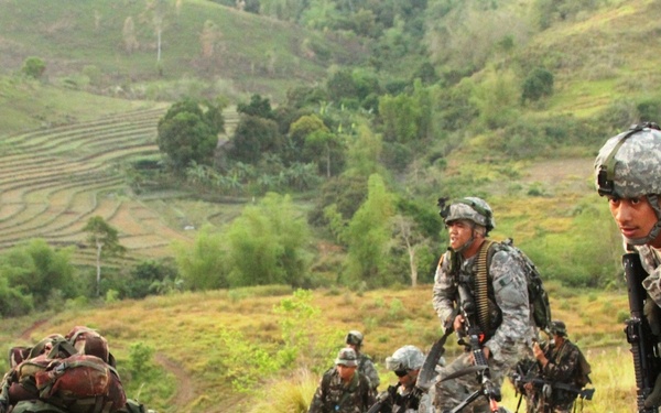 The image shows a joint military training exercise taking place on a hillside terrain with terraced agricultural fields visible in the background. Soldiers in two distinct camouflage patterns — US Army ACU (Army Combat Uniform) and what appears to be Philippine Army camouflage — are conducting a tactical movement drill. The mixed unit composition and non-combat context suggest this is a bilateral military exercise, likely Balikatan or a similar US-Philippines joint training operation.