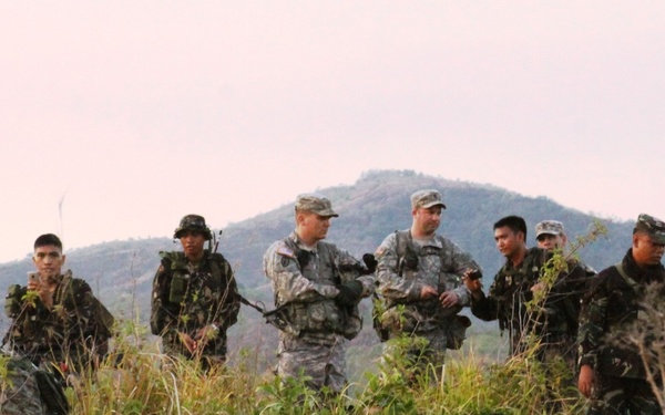 A group of military personnel in camouflage uniforms are gathered on a hillside or elevated terrain covered with tall grass and vegetation. The group appears to include soldiers in US Army ACU (Army Combat Uniform) pattern alongside soldiers in darker camouflage, suggesting a joint or combined military exercise. A mountainous landscape is visible in the background under an overcast sky.