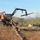 A logging operation is underway in a partially cleared wooded area. A tracked logging machine with a hydraulic grapple arm is lifting a log, while a worker with a chainsaw operates nearby. The site shows extensive tree clearing with numerous felled logs and debris scattered across the ground.