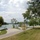A well-maintained lakeside park area featuring paved pathways, picnic facilities, and mature trees along a calm, wide body of water. The scene depicts a recreational day-use area with amenities such as picnic tables, grills, and light poles scattered near the shoreline. The landscape is lush and green, consistent with late summer conditions.