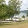 A lakeside park or recreation area photographed from an elevated vantage point on a sunny day. The scene shows a well-maintained grassy slope leading down to a waterfront area with paved paths, picnic facilities, and temporary event tents near the water's edge. A large lake or reservoir stretches into the background, bordered by forested hills.