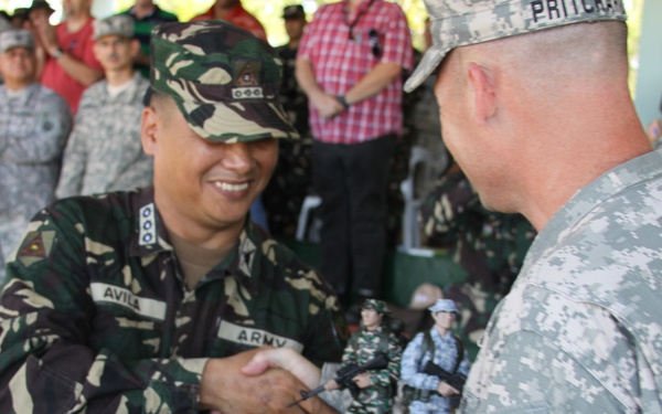 Two military officers are engaged in a formal handshake ceremony, with one wearing Philippine Army camouflage uniform and the other in US Army ACU (Army Combat Uniform). The Philippine Army officer, identified by the name tape 'AVILA,' is smiling while exchanging what appears to be a commemorative plaque or gift with the US Army officer named 'PRITCHARD.' The event appears to be a formal military exchange or ceremony attended by numerous soldiers in the background.