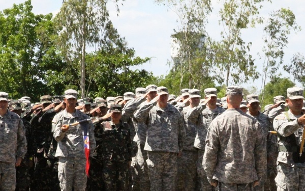 A military formation ceremony is taking place on an outdoor parade ground, featuring soldiers in two distinct uniforms — US Army ACU (Army Combat Uniform) in digital camouflage and what appear to be Philippine Armed Forces soldiers in darker woodland camouflage. The troops are rendering a salute in formation, with a color guard bearing a red and yellow unit guidon/flag visible on the right side. The setting appears to be a joint military exercise or ceremony, likely in the Philippines based on the mixed forces present.