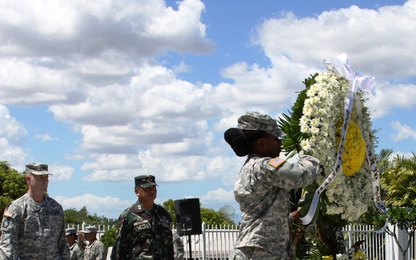 A military wreath-laying ceremony is taking place outdoors, involving personnel in two distinct uniforms — US Army soldiers in ACU (Army Combat Uniform) pattern and what appear to be Philippine Armed Forces soldiers in woodland camouflage. Two soldiers are carrying a large floral wreath with white and yellow flowers toward what is likely a memorial or monument. A formation of troops stands at attention in the background.