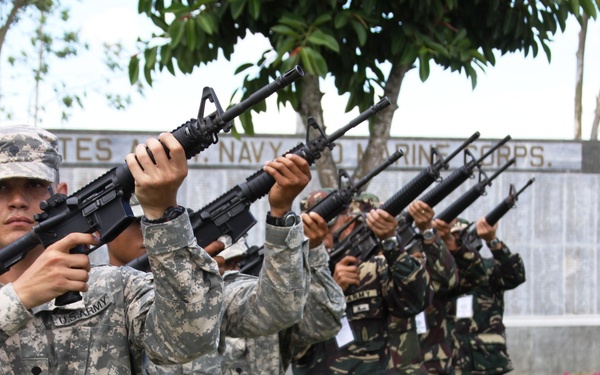 A mixed formation of U.S. Army soldiers and what appear to be Philippine military personnel are performing a rifle salute or ceremonial volley fire. The soldiers are lined up in a row, raising M16-style rifles overhead in unison. A memorial wall with partially visible text referencing 'United States,' 'Navy,' and 'Marine Corps' is visible in the background.