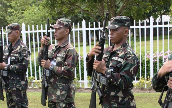 The image shows a military formation or drill exercise taking place on a grass field near a white fence. Several soldiers wearing 'ARMY' labeled camouflage uniforms are holding rifles in a ceremonial or training stance. At least one soldier in US Army ACU (Army Combat Uniform) pattern is also visible on the left side, suggesting a joint or combined military exercise.
