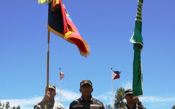 A formal military ceremony featuring soldiers from two distinct forces: US Army personnel in ACU (Army Combat Uniform) digital camouflage on the left, and what appears to be Philippine Army soldiers in woodland camouflage on the right. Two color guards are bearing military guidons/flags, with an American flag and Philippine flag visible on flagpoles in the background. The central figure wearing a name tape reading 'LEONES' and 'ARMY' tab appears to be a Philippine Army officer presiding over the ceremony.