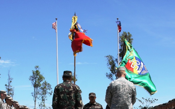 A formal military ceremony is taking place at what appears to be a memorial or ceremonial grounds, featuring soldiers from two different nations standing on designated platforms labeled 'HOST' and 'HONOREE'. Two color guard bearers hold military flags/guidons while formations of soldiers in different uniforms are lined up on both sides. The setting includes white gate pillars, flagpoles with flags, and floral arrangements, suggesting a joint military commemoration event.