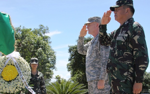 A formal military ceremony showing two senior officers — one in US Army ACU (Army Combat Uniform) and one in Philippine Army camouflage — rendering salutes together on a raised platform. A floral wreath with ribbons is prominently displayed in the background, suggesting a memorial or commemorative event. Additional military personnel from both forces are visible in attendance.