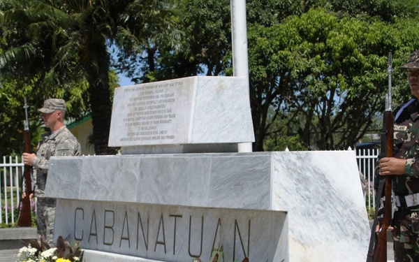 A memorial ceremony is taking place at the Cabanatuan memorial monument in the Philippines. Two soldiers — one in U.S. Army combat uniform and one in Philippine Army camouflage — stand at ceremonial guard positions flanking a white marble monument. Floral wreaths have been placed at the base of the monument in tribute.