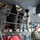 Military maintenance personnel are performing aircraft maintenance work inside a hangar. Workers on an elevated metal scaffold platform are accessing open panels on the underside of a large military aircraft. One airman at ground level appears to be operating a laptop or control device.