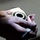 A pair of hands is holding a small metallic gear or splined mechanical component, appearing to clean or polish it with a pink abrasive cloth or sandpaper. The component has visible internal splines or teeth and a hollow center, suggesting it is a mechanical coupling or hub. The image is a close-up shot with shallow depth of field, emphasizing the hands and component.