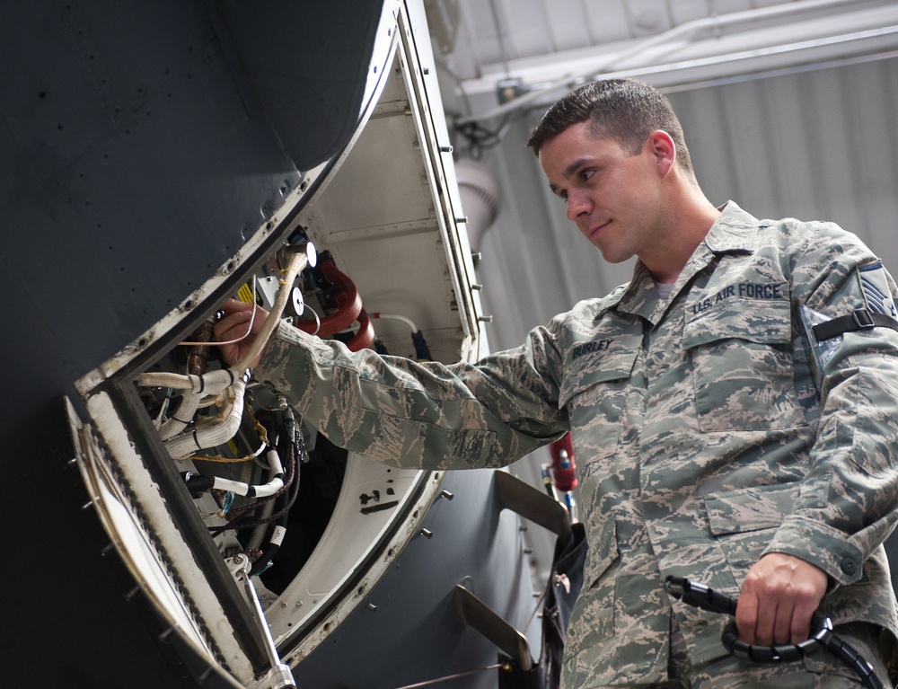 A U.S. Air Force airman in ABU (Airman Battle Uniform) camouflage is performing maintenance on a military aircraft inside a hangar. The technician is inspecting or working on an open access panel that reveals internal wiring, cables, and components. The individual's name tape and 'U.S. AIR FORCE' tape are visible on the uniform.
