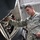 A U.S. Air Force airman in ABU (Airman Battle Uniform) camouflage is performing maintenance on a military aircraft inside a hangar. The technician is inspecting or working on an open access panel that reveals internal wiring, cables, and components. The individual's name tape and 'U.S. AIR FORCE' tape are visible on the uniform.