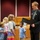 A formally dressed adult male is reading from a black folder/document in what appears to be a government chamber or courtroom setting. Four young children of varying ages stand before him, appearing to listen attentively. The setting and formal document suggest an official ceremony, possibly a proclamation reading or recognition event.