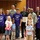 A group photo of adults and children gathered for what appears to be a military family recognition ceremony. Several individuals are wearing purple 'Purple Up! For Military Kids' t-shirts, and one person is holding a framed award or plaque. Two individuals in military uniforms (U.S. Army and U.S. Air Force) are present among the group.