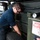 A mechanic wearing safety glasses and dark blue coveralls is working on a military-style vehicle, appearing to inspect or service the wheel/tire area. The vehicle is olive drab green and bears a partially visible label reading 'HYDRAULIC FLUID' along with a 'DANGER' warning sticker. The work appears to be taking place in a maintenance bay or garage setting.