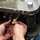 A person is manually threading a nut onto a bolt near a large piece of industrial or heavy machinery. The machinery appears to be dark green/black painted metal with multiple bolts and a large cylindrical component. The work appears to be assembly or maintenance of mechanical equipment.