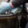 A person wearing light blue nitrile gloves and dark clothing is working on top of a large industrial metal drum or barrel. The drum appears heavily worn with rust, oil stains, and paint deterioration. The worker appears to be manipulating or tightening a central bolt or fitting on the drum's lid.