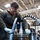 A male technician or mechanic is working on industrial equipment in a large workshop or maintenance facility. He is wearing protective safety goggles and blue rubber gloves while handling blue-and-black striped hydraulic or pneumatic hoses connected to a heavy metal component. The setting appears to be a military or industrial maintenance bay based on the equipment and attire visible.