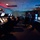 A dimly lit military or air traffic control operations center with multiple workstations featuring radar and computer displays. Personnel in military uniform and civilian clothing are present, with some seated at consoles and two individuals standing and conversing. An American flag is visible on the right wall, and a digital clock display showing '15:04' is mounted on the left wall.