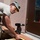 A person wearing military camouflage cap and yellow safety glasses is performing carpentry work near a building entrance. They are working with wooden boards and a cordless power drill, appearing to construct or repair a wooden structure such as steps or a ramp adjacent to a door. The setting is outdoors against a stucco wall with a brown door visible.
