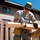 A man wearing military-style camouflage pants, a gray t-shirt, and a camouflage cap is using a circular saw to cut a wooden board outdoors. Sawdust and wood particles are visibly flying through the air during the cutting operation. He is working near a building, with wooden fence boards and a sawhorse visible in the foreground.