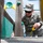 A U.S. Air Force service member named 'Maher' is working on a wooden playground structure, using a tape measure to take measurements. The individual is wearing the standard Air Force camouflage uniform (ABU - Airman Battle Uniform) with a patrol cap and sunglasses. The setting appears to be an outdoor playground, suggesting a community service or base improvement project.