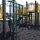 A person in military camouflage uniform is inspecting or documenting something on a playground structure, appearing to write on a clipboard or form. The playground features yellow and green equipment including slides, climbing structures, and circular windows, set on black rubber mulch surfacing. The setting appears to be on a military base, visible by the open grounds and institutional buildings in the background.