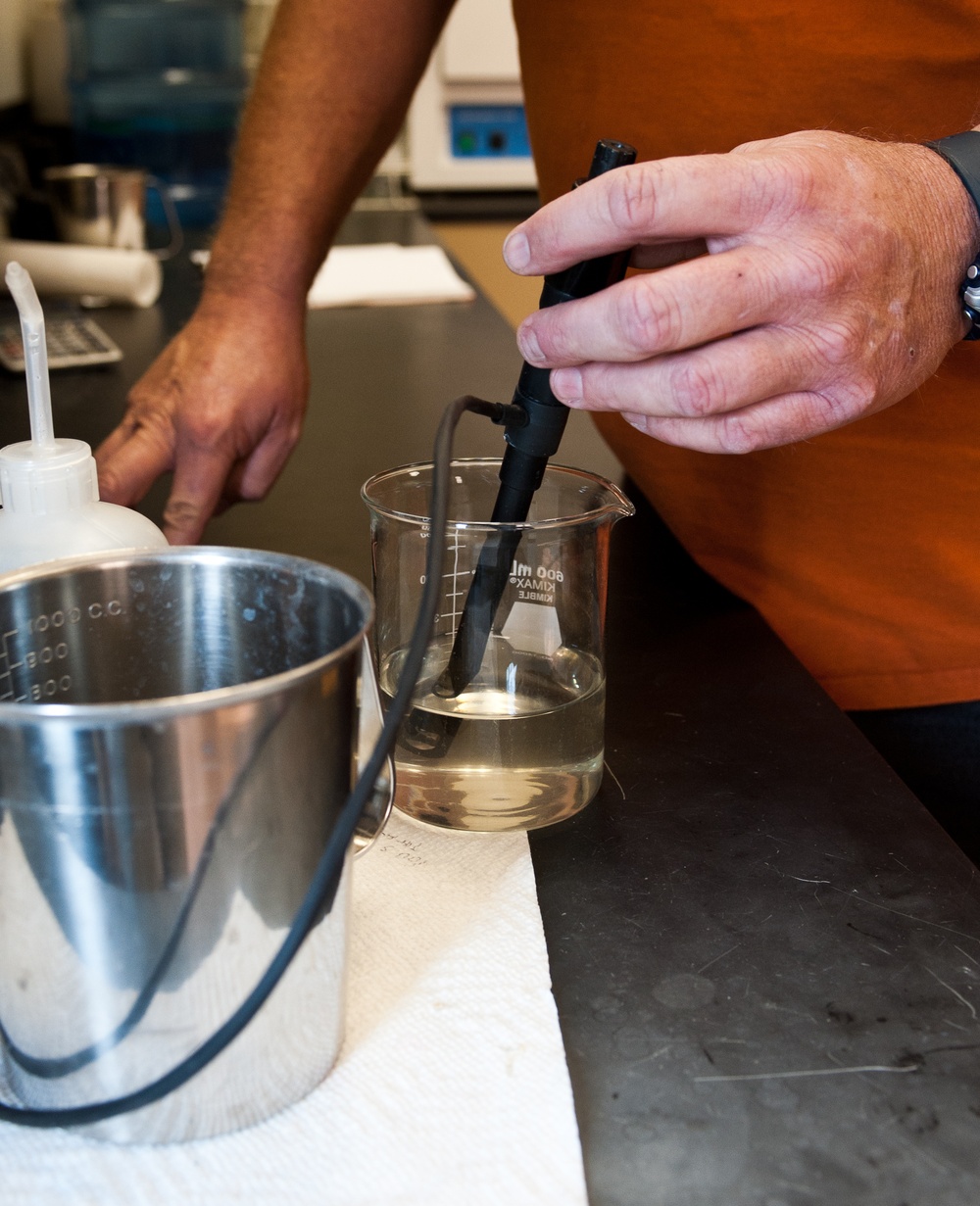 A person wearing an orange shirt is conducting a laboratory procedure, inserting what appears to be a probe or electrode into a glass beaker containing a clear liquid. The beaker is branded 'KIMAX KIMBLE' with a 600 mL capacity marking. The setting is a laboratory workbench with various equipment visible in the background.