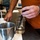A person wearing an orange shirt is conducting a laboratory procedure, inserting what appears to be a probe or electrode into a glass beaker containing a clear liquid. The beaker is branded 'KIMAX KIMBLE' with a 600 mL capacity marking. The setting is a laboratory workbench with various equipment visible in the background.