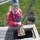 An older man with a white beard, wearing denim overalls and a red long-sleeve shirt, is cleaning or clearing debris from a storm drain or catch basin using a long-handled tool. He is lifting a clump of compacted sediment or mud from the open drain compartment. A metal cylindrical container nearby appears to be collecting the removed material.