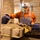 A middle-aged man wearing denim overalls and an orange long-sleeve shirt works with industrial machinery in what appears to be a water treatment or waste processing facility. He is inspecting or operating equipment that appears to be processing sludge or debris material on a conveyor or press mechanism. The setting is an industrial interior with concrete block walls.