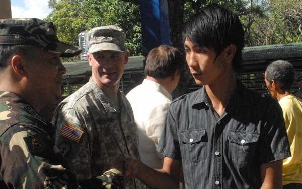 A military ceremony or distribution event showing a soldier in camouflage uniform handing a document to a young civilian. A U.S. Army soldier (identified by American flag patch) stands nearby smiling, suggesting a joint military cooperation event. The setting appears to be an outdoor covered area in a tropical location, likely the Philippines based on the Filipino military camouflage pattern.