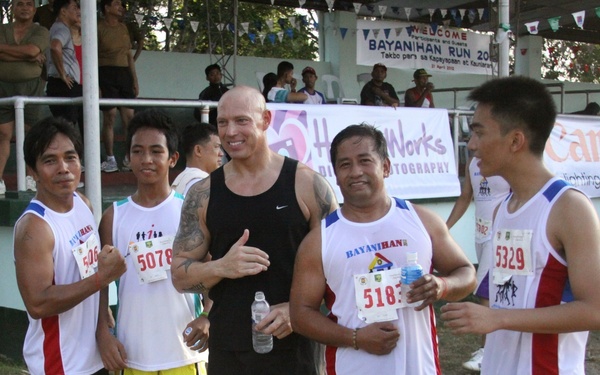 A group of five men pose together at what appears to be the 'Bayanihan Run 2012' running event. Four of the men are wearing race bibs with numbers (506, 5078, 5183, 5329), while one taller man in a black Nike tank top stands in the center without a bib. The event appears to be held at an outdoor venue with bleachers and sponsor banners visible in the background.