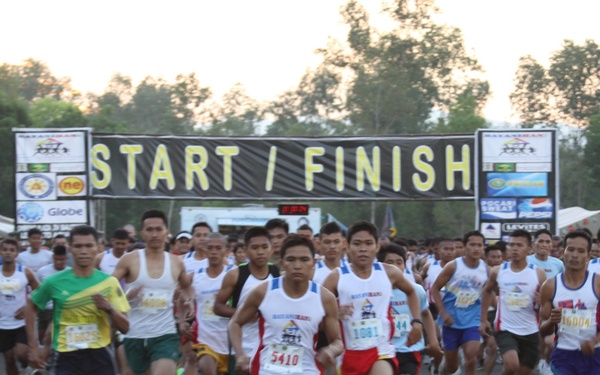 A large group of runners is crossing or departing from a START/FINISH line at an outdoor road race event. The scene appears to be taken at dawn or early morning, with trees visible in the background and tents set up along the sides. Participants are wearing numbered race bibs and athletic clothing.