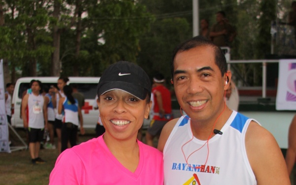 Two runners pose together at what appears to be a road race or fun run event. The woman on the left wears bib number 16043 and a pink 'Wahine Half Marathon' shirt with a Nike cap, while the man on the right wears bib number 16028 and a 'Bayanihan' race singlet with earphones. The background shows other participants, a Red Cross vehicle, and event banners in an outdoor setting.