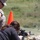 A civilian woman in a black shirt is firing a rifle in the prone position at what appears to be a military shooting range. A male service member in MARPAT camouflage uniform is supervising and observing her technique. A spent brass casing is visibly ejecting from the rifle, indicating the weapon has just been fired.