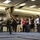 A military personnel in camouflage uniform is being thrown or performing a martial arts/combat technique on blue floor mats in what appears to be an indoor training facility. A group of civilian women wearing lanyards/badges are watching the demonstration from the perimeter. The setting appears to be a formal demonstration or training event, likely a military outreach or orientation program.