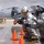 Two firefighters in full aluminized proximity suits are operating a fire hose on what appears to be a military airfield tarmac during a training exercise. Orange traffic cones are positioned in front of them as markers, and military helicopters are visible in the background along with several personnel in military uniforms. The setting and equipment suggest this is a crash/fire/rescue (CFR) training drill at a military aviation installation.