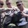 Three U.S. Marines in desert MARPAT camouflage uniforms are gathered outdoors, smiling and examining a military flight helmet. Two Marines are handling the olive-green helmet together while a third observes from behind. The setting appears to be an open field or training area in daylight.