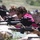 A group of individuals are lying in a prone shooting position at an outdoor firing range, each aiming AR-style rifles downrange. The shooters are using sandbags as rests and are equipped with optical sights and safety glasses. The setting appears to be a supervised marksmanship training or recreational shooting event in a rural, open-air environment.