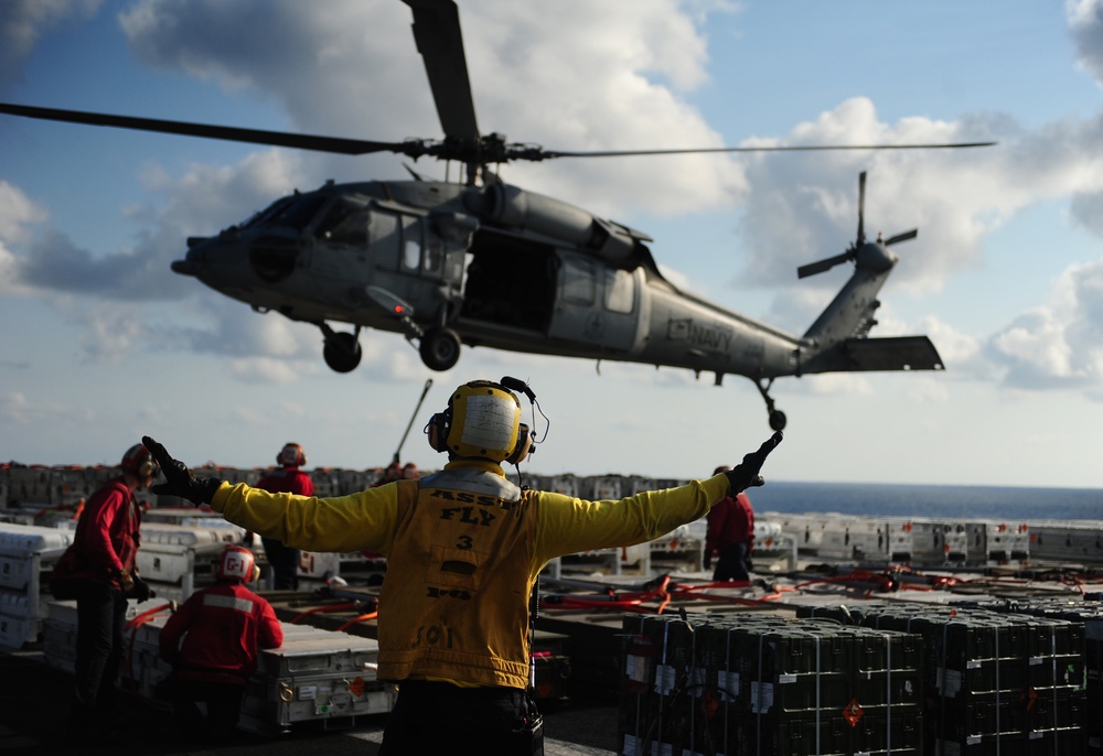 A U.S. Navy flight deck crew member in a yellow jersey is directing an MH-60 Seahawk helicopter during flight operations aboard an aircraft carrier. The deck is loaded with stacked cargo containers and equipment, with additional red-jersied crew members visible in the background. The scene depicts an active underway replenishment or vertical replenishment (VERTREP) operation at sea.