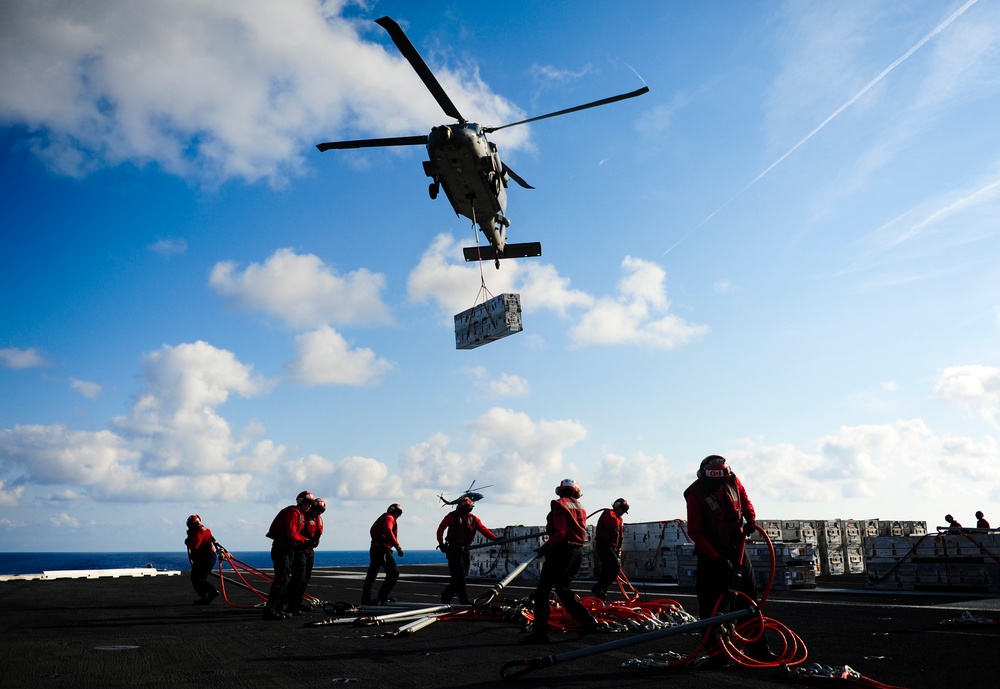A military helicopter performs a vertical replenishment (VERTREP) operation over the deck of a naval vessel at sea, lowering a cargo pallet via sling load. Multiple crew members dressed in red flight deck jerseys and safety gear are positioned on the deck managing lines and receiving the cargo. The scene takes place under a partly cloudy blue sky with the ocean visible in the background.